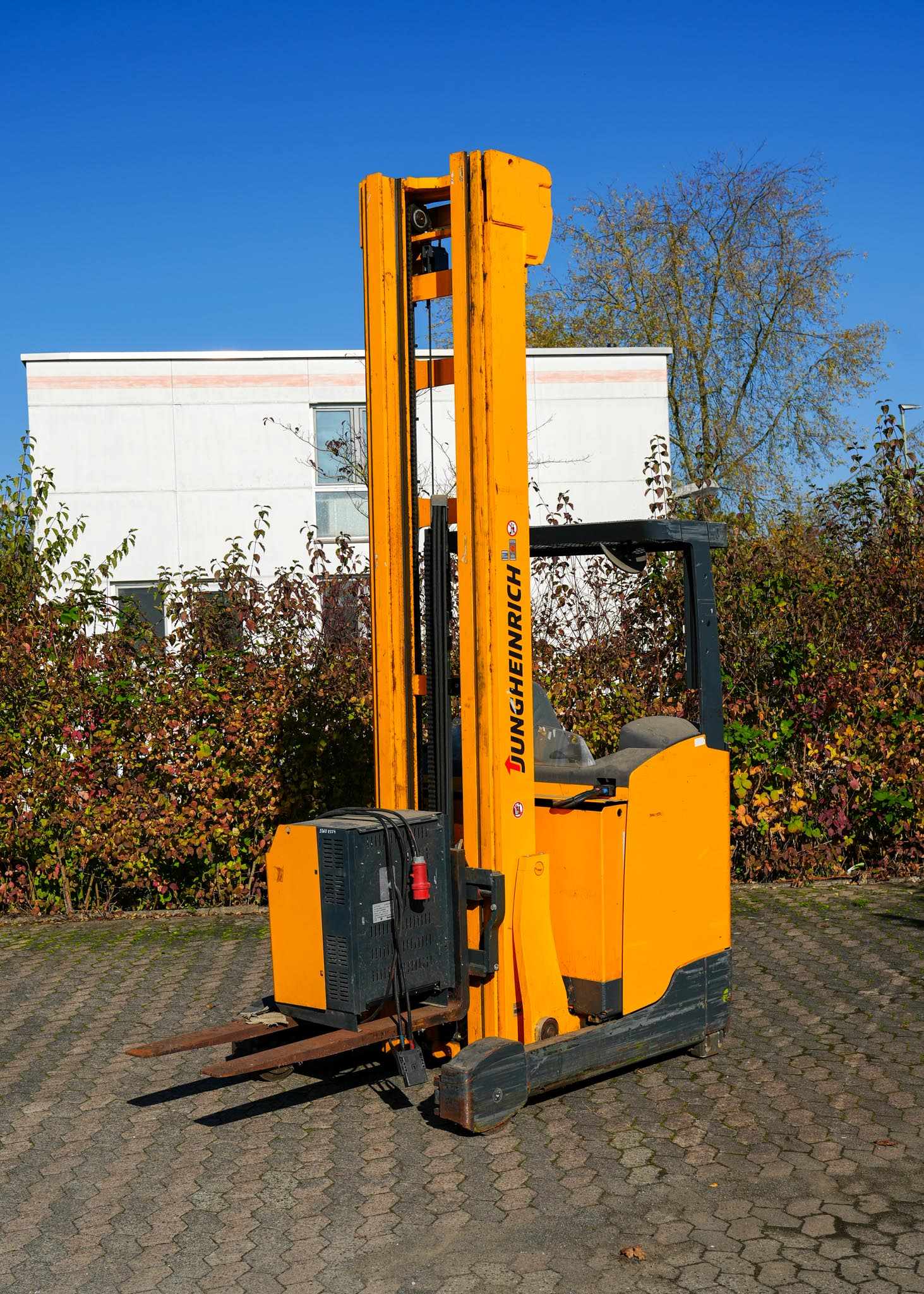 Ein gelber Gabelstapler vor einem Gebäude auf gepflastertem Boden, umgeben von Herbstlaub.