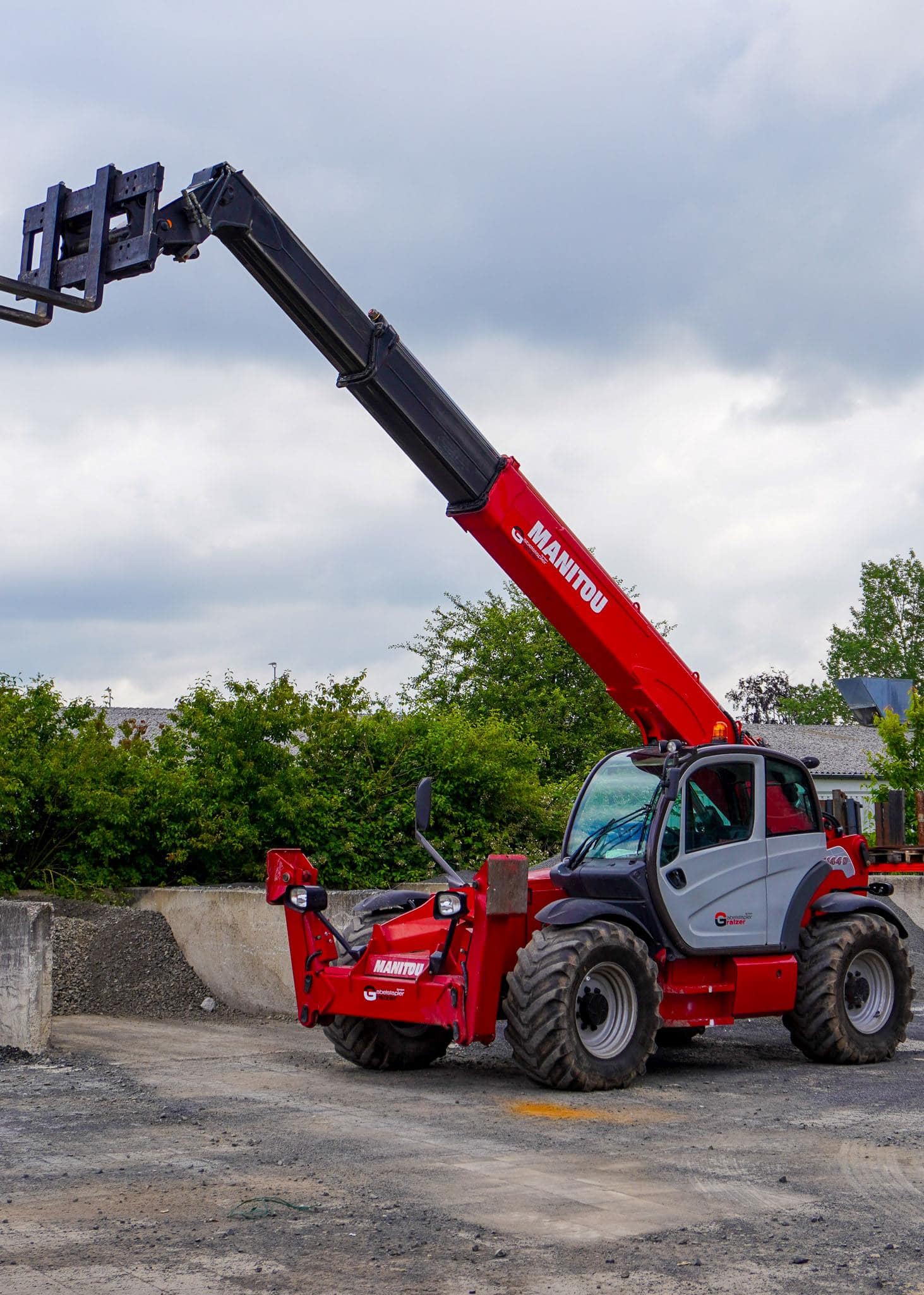 Ein roter Teleskoplader mit ausgefahrenem Arm steht auf einer Baustelle unter bewölktem Himmel.