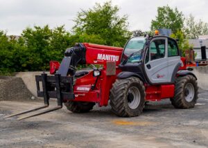 Roter Teleskopstapler auf Baustelle, graue Kabine, abgestellt auf Kies, umgeben von Bäumen.