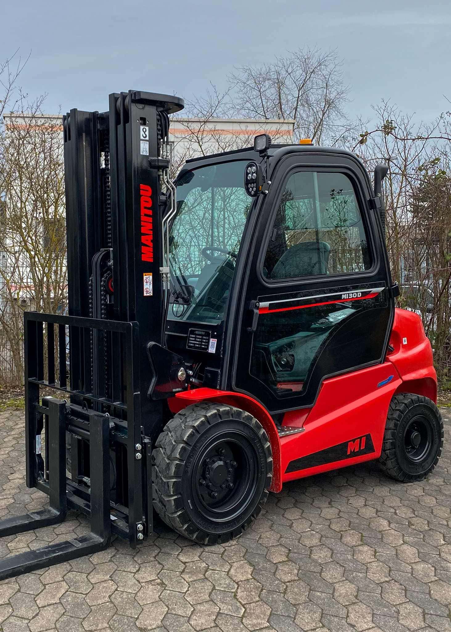 Ein roter Manitou-Gabelstapler steht auf einem gepflasterten Platz vor kahlen Bäumen.