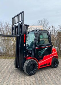 Ein roter Gabelstapler, Modell "Manitou", parkt auf einer gepflasterten Fläche vor Büschen.